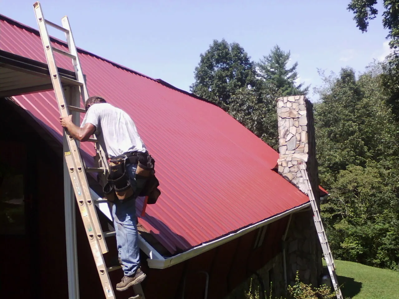 Roofer on a ladder beside a red metal roof and stone chimney — Soffit & Fascia Repair by Roofing Brothers in Asbury